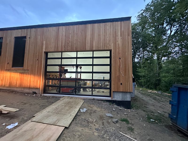 Contemporary full-view glass garage door on cedar building at dusk with interior lit — MA